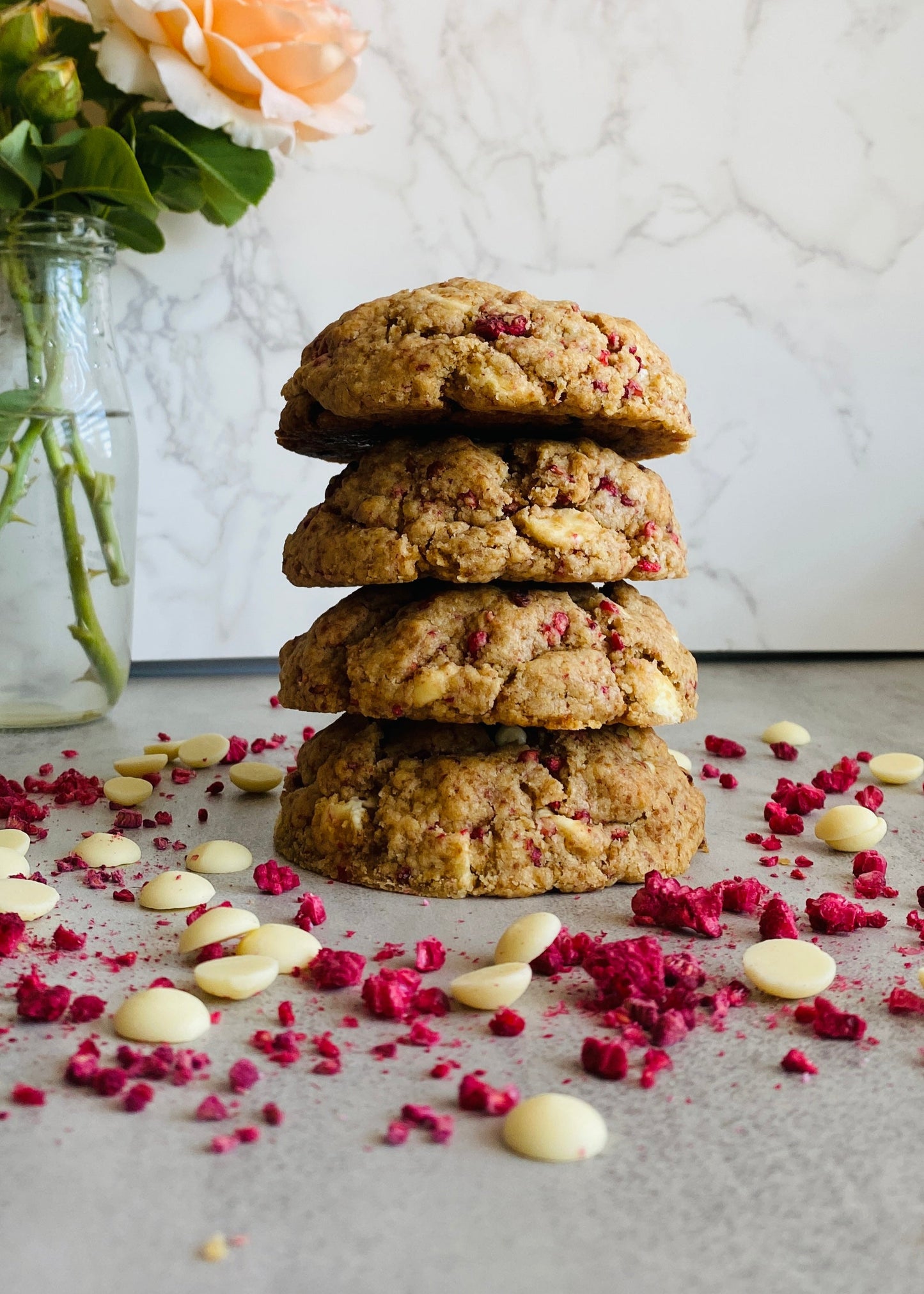 Stack of cookies with white chocolate chips and freeze-dried raspberries on a marble surface with flowers in the background.