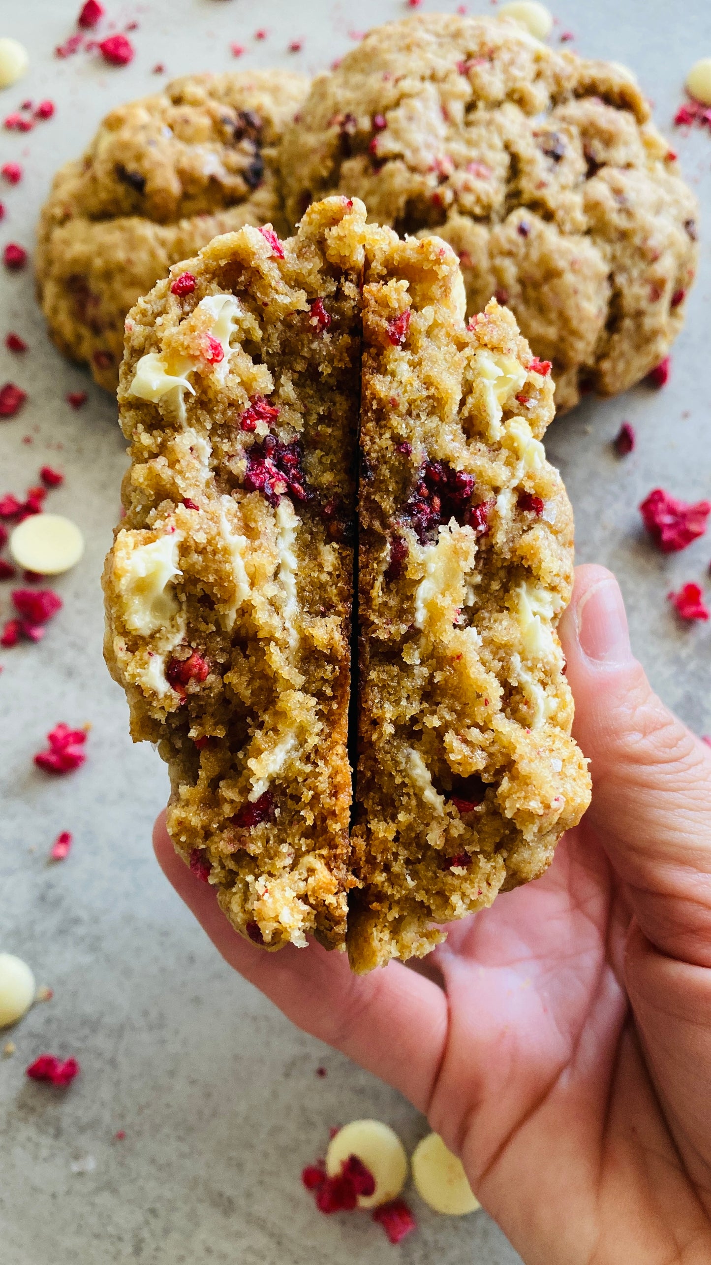 Hand holding a cookie with a bite taken out, showing a crumbly interior, on a surface with cookies and raspberries and white chocolate chips.