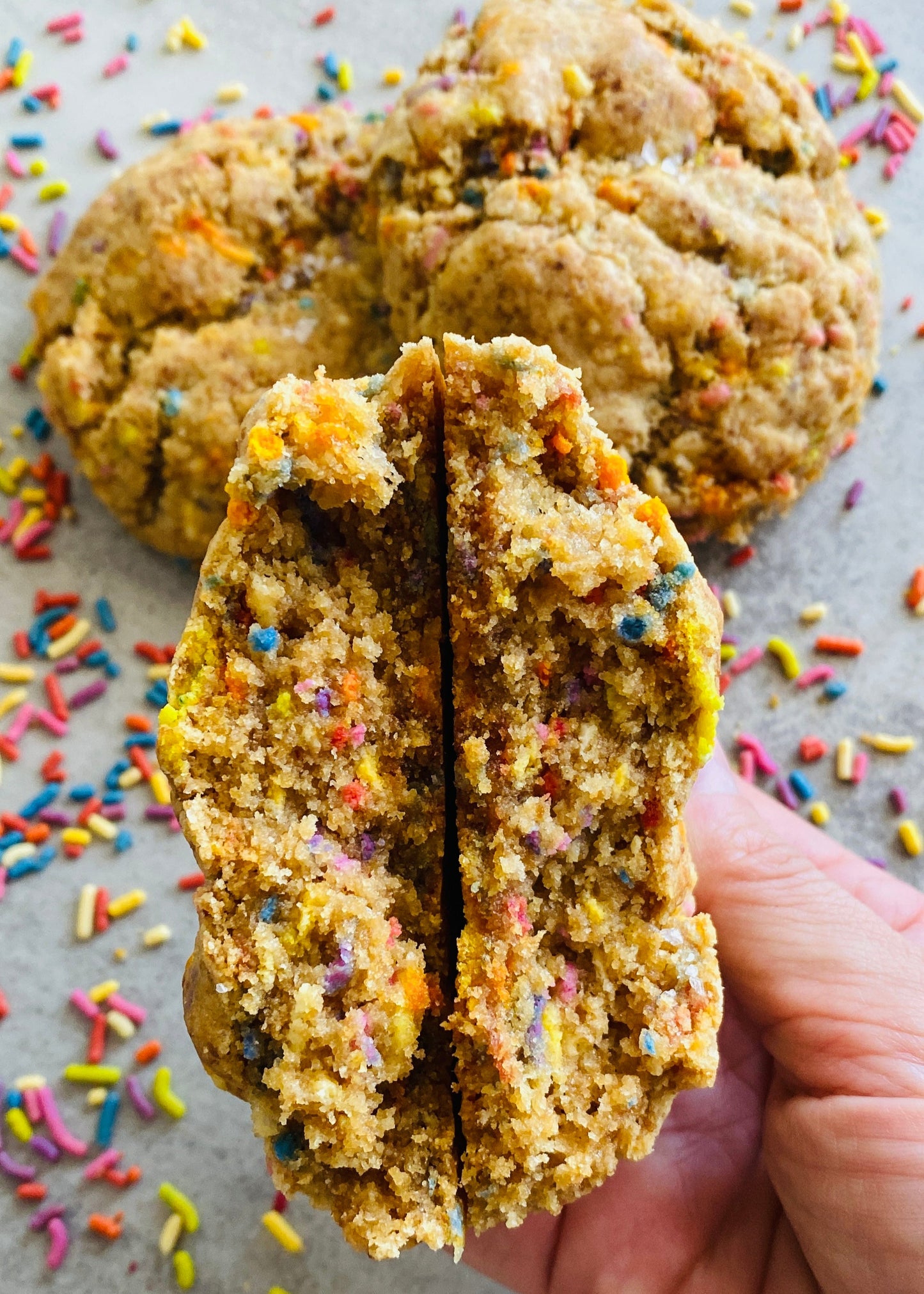 Hand holding a split oatmeal cookie with colorful sprinkles on a gray surface