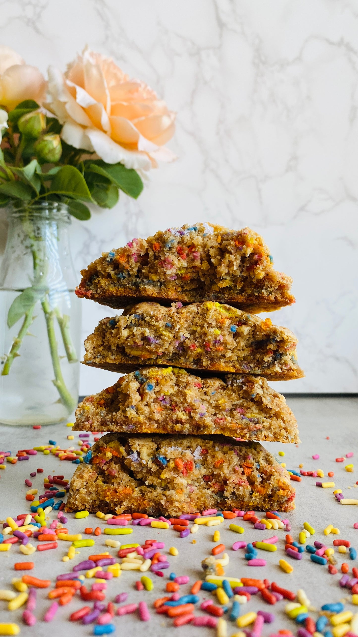 Stack of colorful cookies with sprinkles on a light surface, next to a vase with flowers.