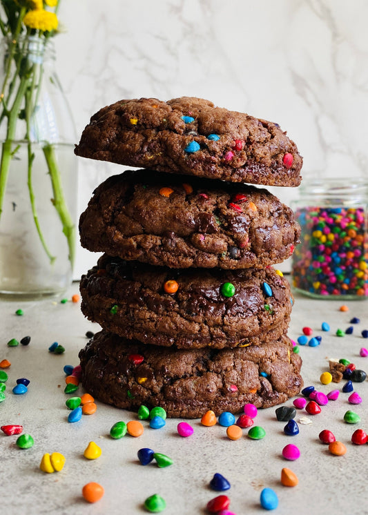 A stack of three cosmic brownie cookies with rainbow sprinkles on top, placed on a light-colored surface with more sprinkles scattered around.