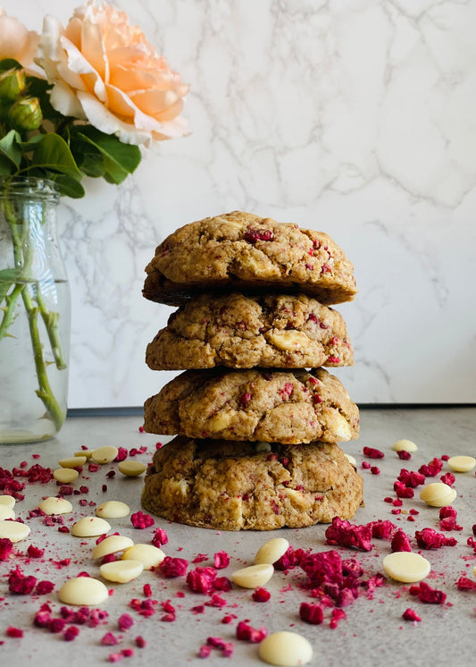 Stack of cookies with white chocolate chips and freeze-dried raspberries on a marble surface with flowers in the background.