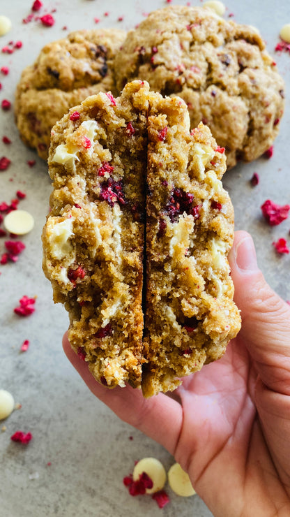 Hand holding a cookie with a bite taken out, showing a crumbly interior, on a surface with cookies and raspberries and white chocolate chips. 